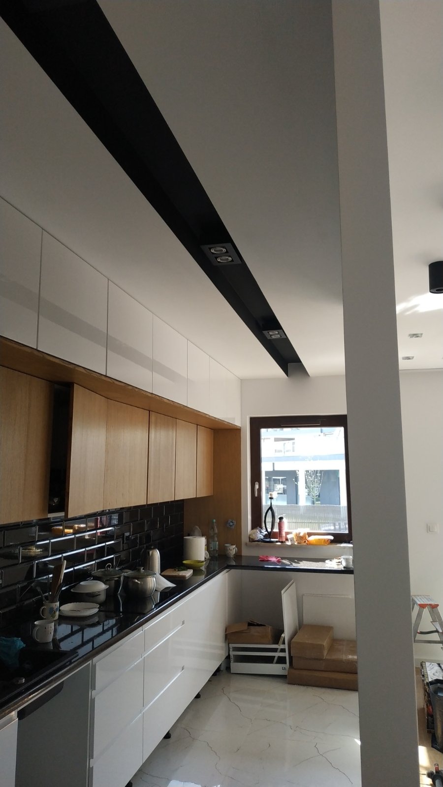 Nowoczesna kuchnia z białymi i drewnianymi szafkami, czarnym blatem i błyszczącymi czarnymi płytkami na ścianie, widoczne czarne oświetlenie sufitowe i marmurowa podłoga.