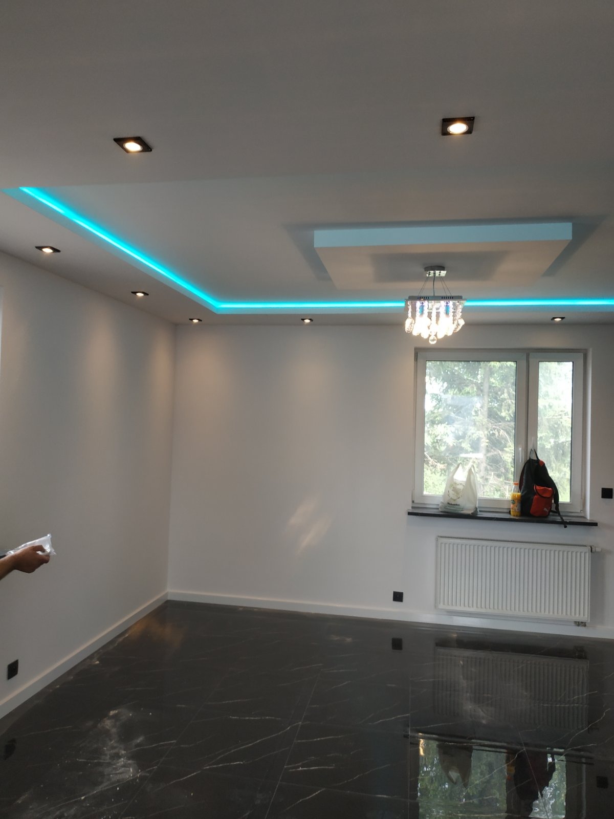 Pusty pokój po remoncie z czarną marmurową podłogą, białymi ścianami, oświetleniem LED na suficie i ozdobnym żyrandolem, torba i plecak leżą na parapecie okna.