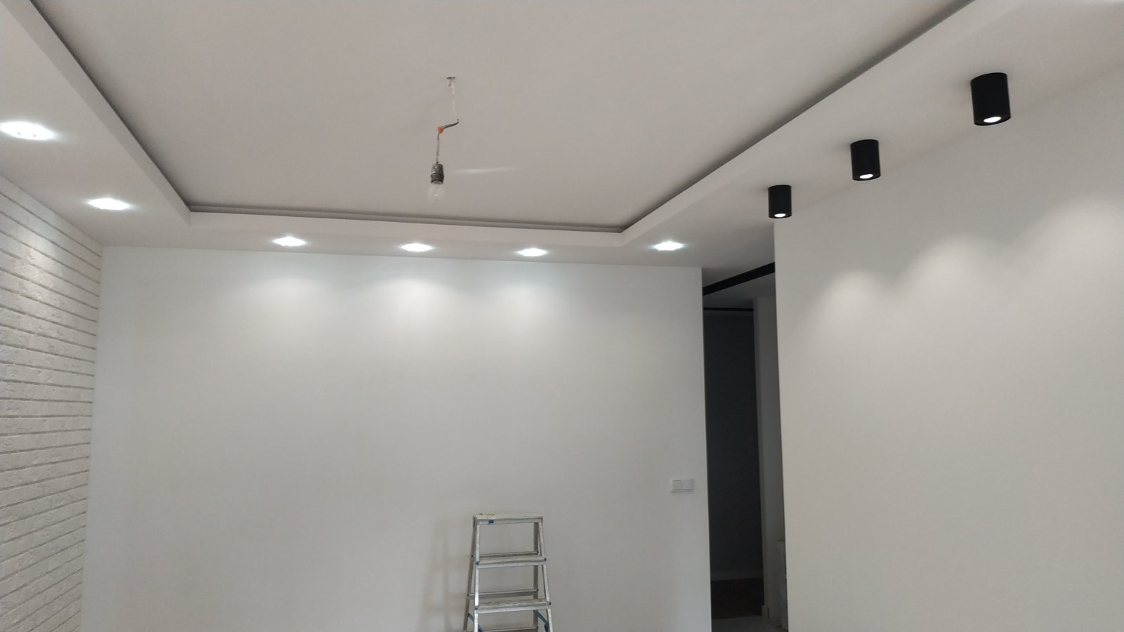 Wnętrze pokoju z białymi ścianami, oświetleniem sufitowym typu downlight i listwą LED, drabina aluminiowa w rogu, pojedyncza żarówka zwisająca z sufitu.