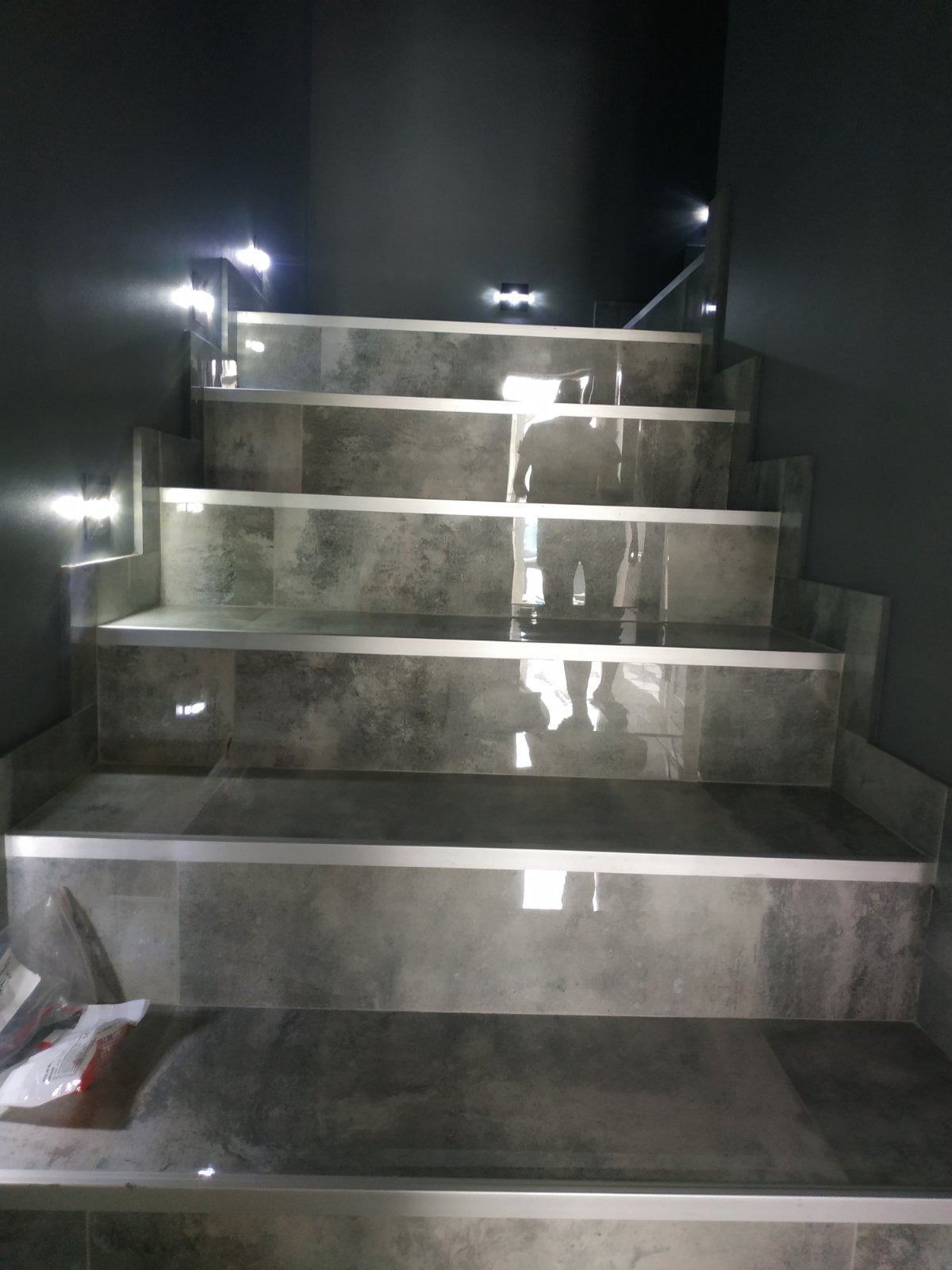 Wykończone schody wewnętrzne z szarymi płytkami imitującymi beton, z aluminiowymi listwami zabezpieczającymi krawędzie, oświetlone punktowymi lampami na ścianie.