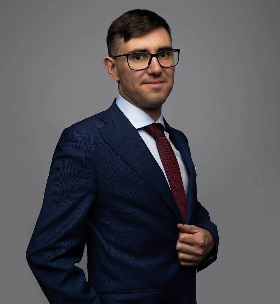 Portret mężczyzny w okularach, w granatowym garniturze i bordowym krawacie, pozującego na szarym tle. Uśmiech, pewny wzrok.