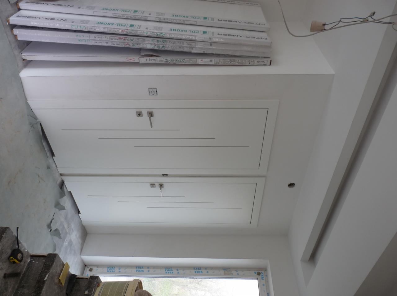 Białe, nowoczesne drzwi rewizyjne w suficie podwieszanym z płyt kartonowo-gipsowych, widoczne opakowania płyt INTER AMBER POL-SKONE, fragment okna z profilem VEKA oraz narzędzia na podłodze.