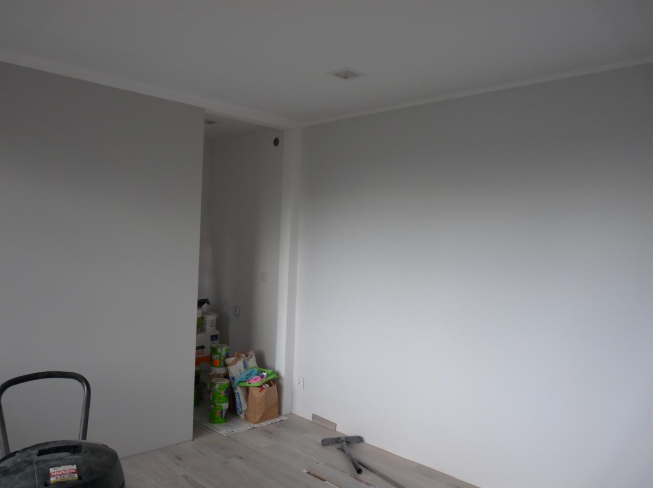 Wnętrze pokoju po malowaniu ścian na szaro i biało, widoczne puszki z farbą, wałek malarski i odkurzacz przemysłowy.