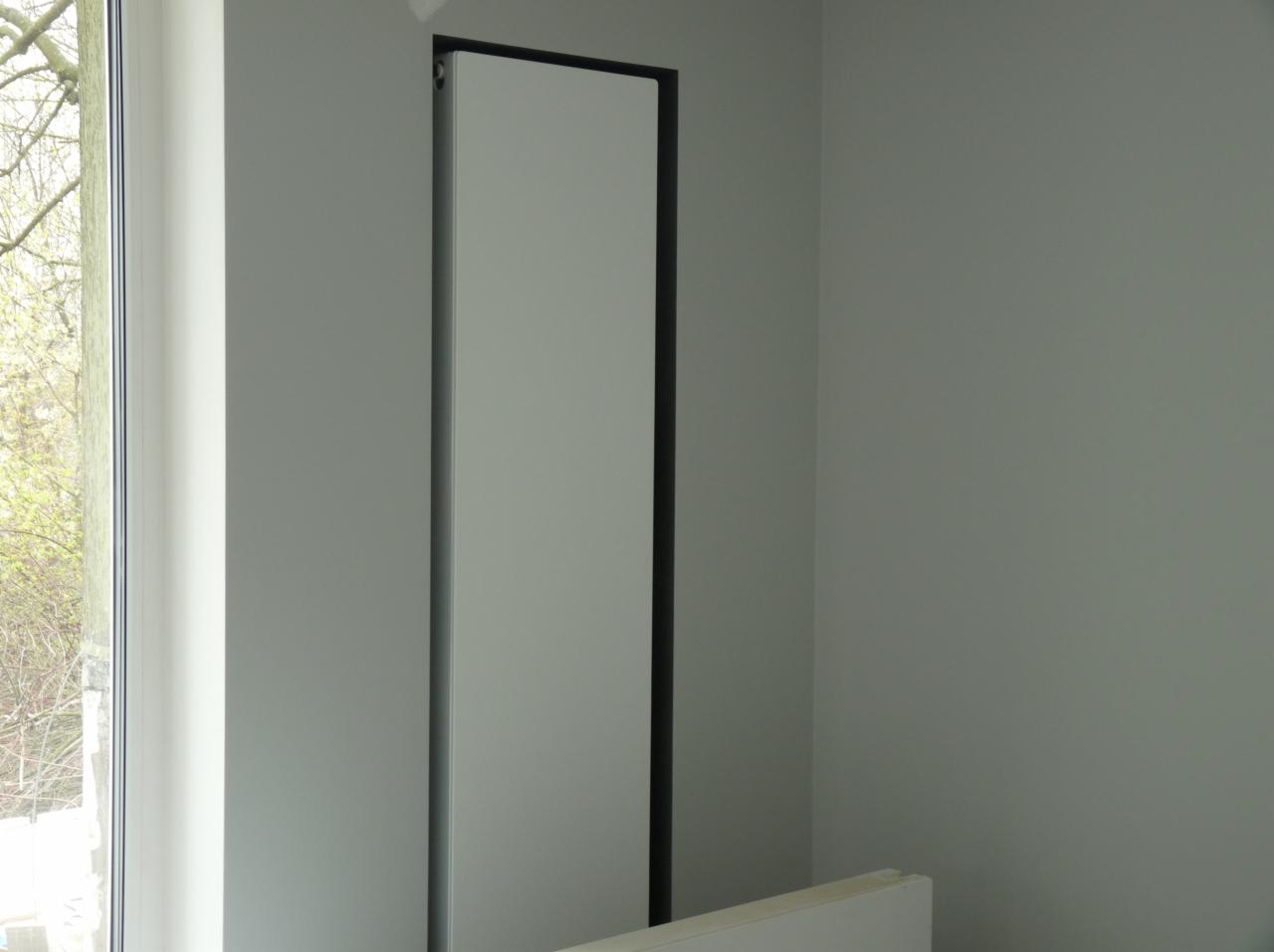 Zamontowana, pionowa płyta kartonowo-gipsowa z czarną ramą przy oknie w szarym pomieszczeniu, widoczne drzewa za oknem.