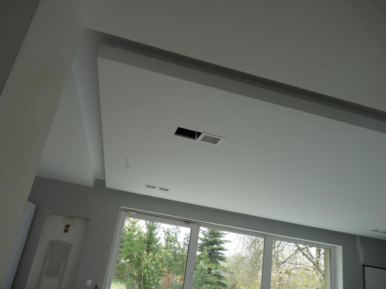 Sufit podwieszany z widocznymi otworami wentylacyjnymi i fragmentem okna z widokiem na zieleń.