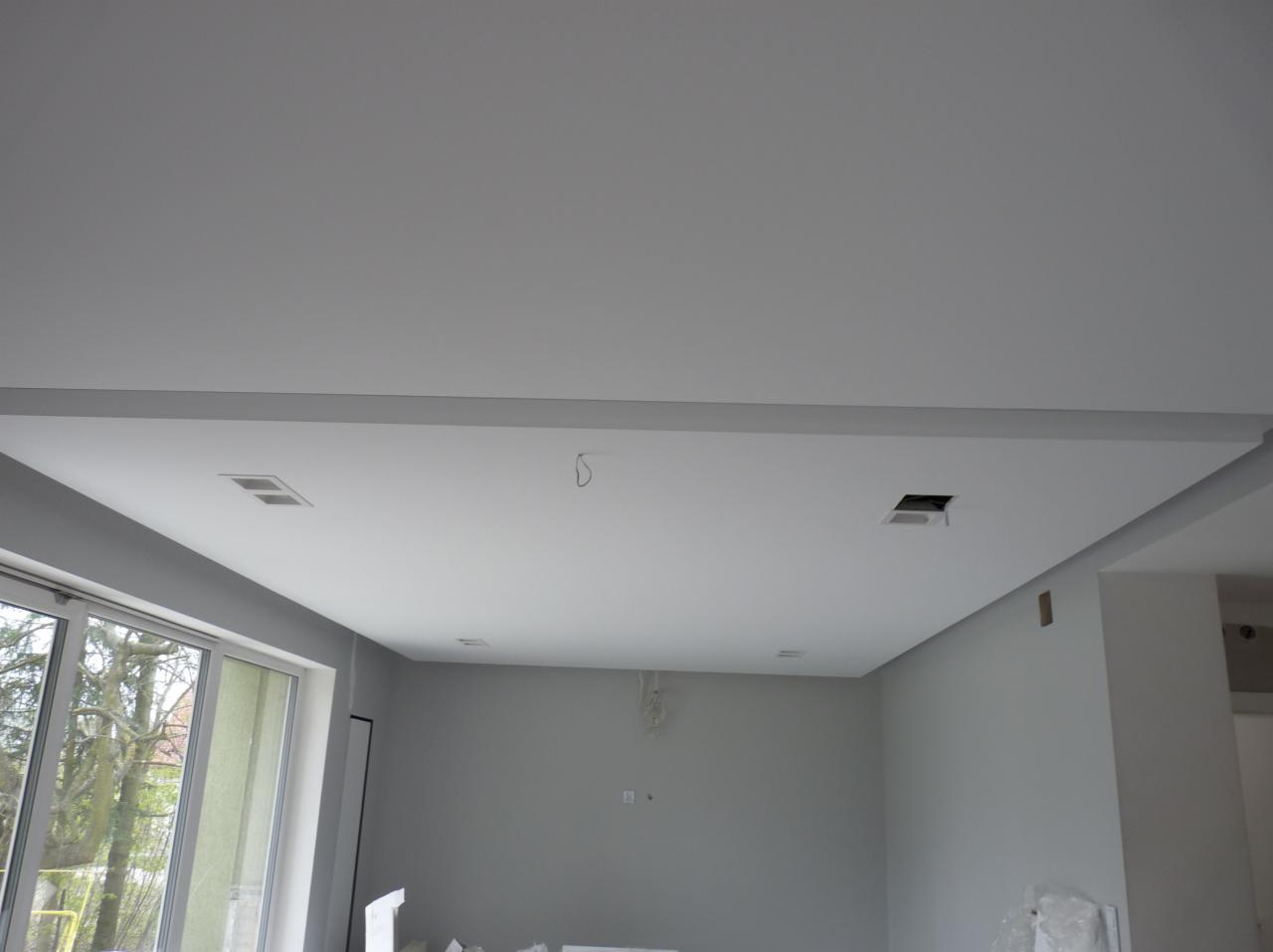 Wykończone wnętrze z gładką, białą powierzchnią sufitu podwieszanego z widocznymi otworami na oświetlenie i elementami instalacji elektrycznej, szara ściana i okno z widokiem na drzewa.