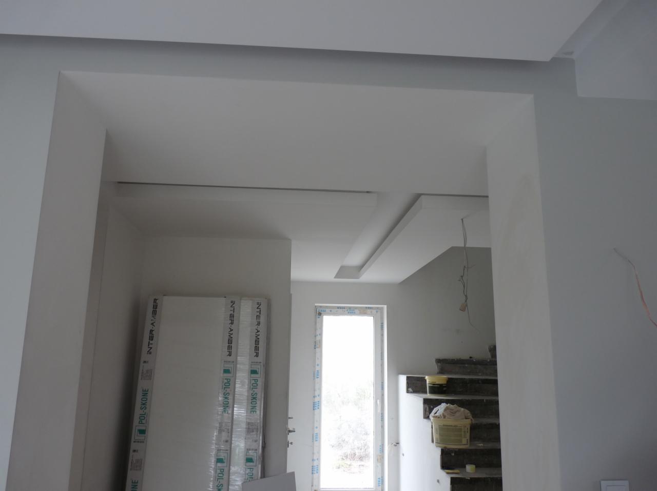 Wnętrze w trakcie wykańczania: białe ściany, sufit z elementami dekoracyjnymi, okno, schody z wiadrem, oraz paczki płyt gipsowo-kartonowych w folii ochronnej opartych o ścianę.