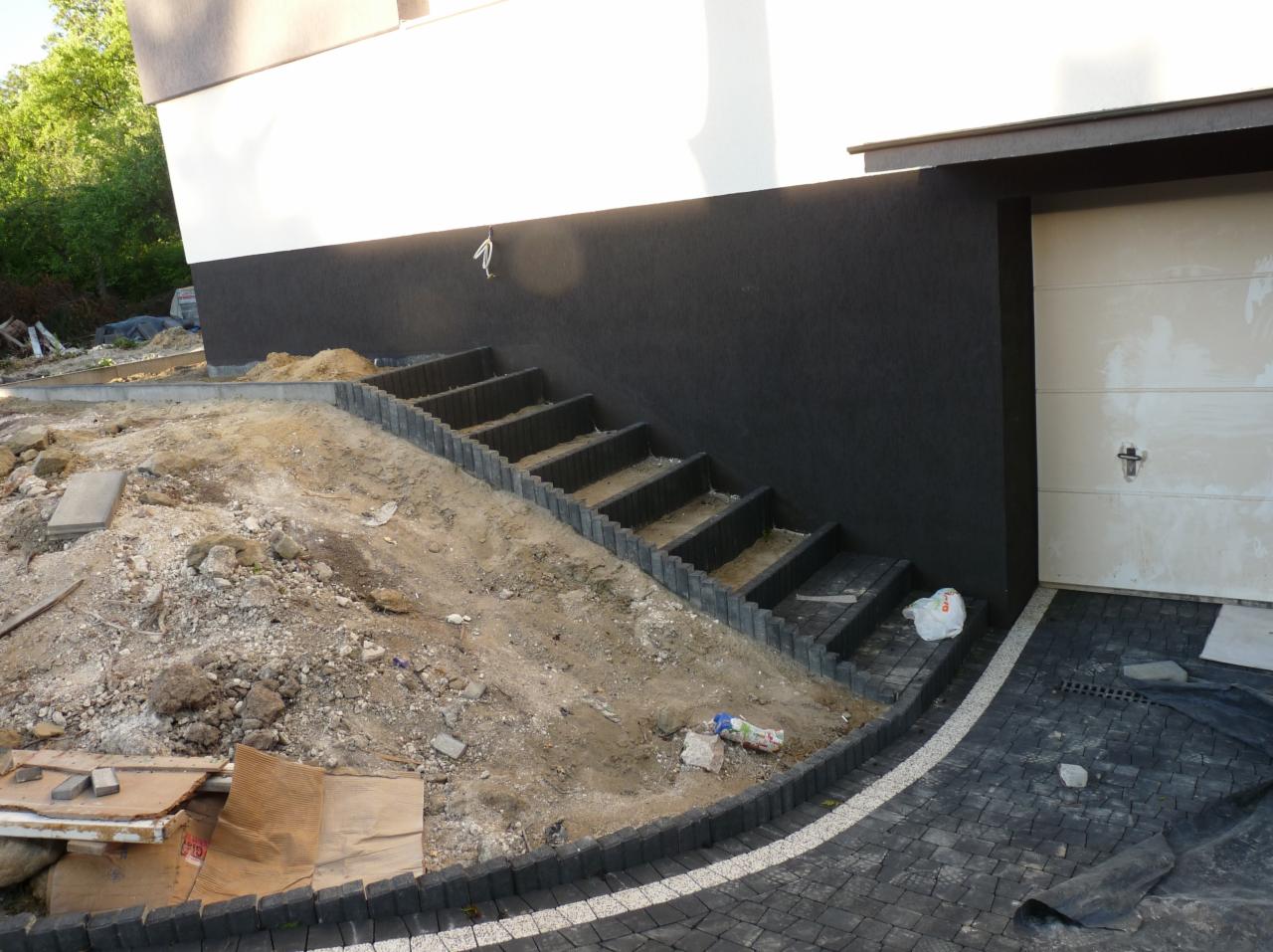 Ukończone schody zewnętrzne z kostki brukowej prowadzące do garażu, widoczny teren po robotach ziemnych z resztkami materiałów budowlanych.