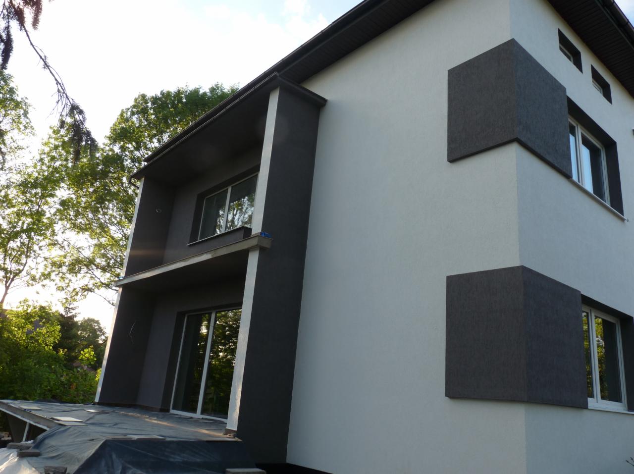 Dwukondygnacyjny dom z elewacją w odcieniach szarości i bieli, z wyraźnie zaznaczonymi, ciemnoszarymi elementami dekoracyjnymi w kształcie prostopadłościanów, widok z dołu na częściowo osłonięty...