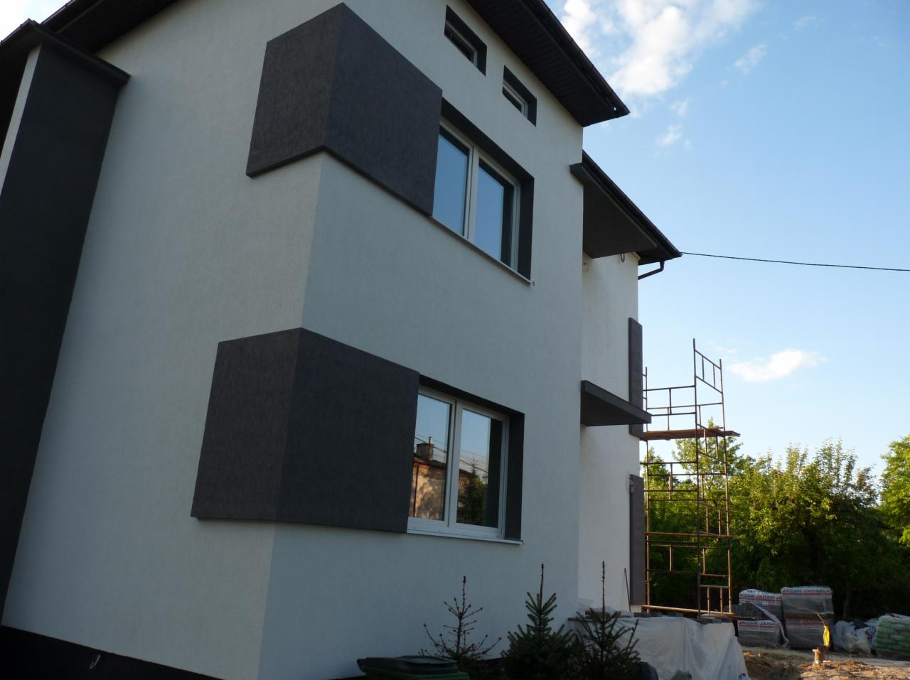 Dwukondygnacyjny dom z elewacją w odcieniach bieli i grafitu, z widocznymi rusztowaniami i materiałami budowlanymi, sugerujący trwające prace wykończeniowe.