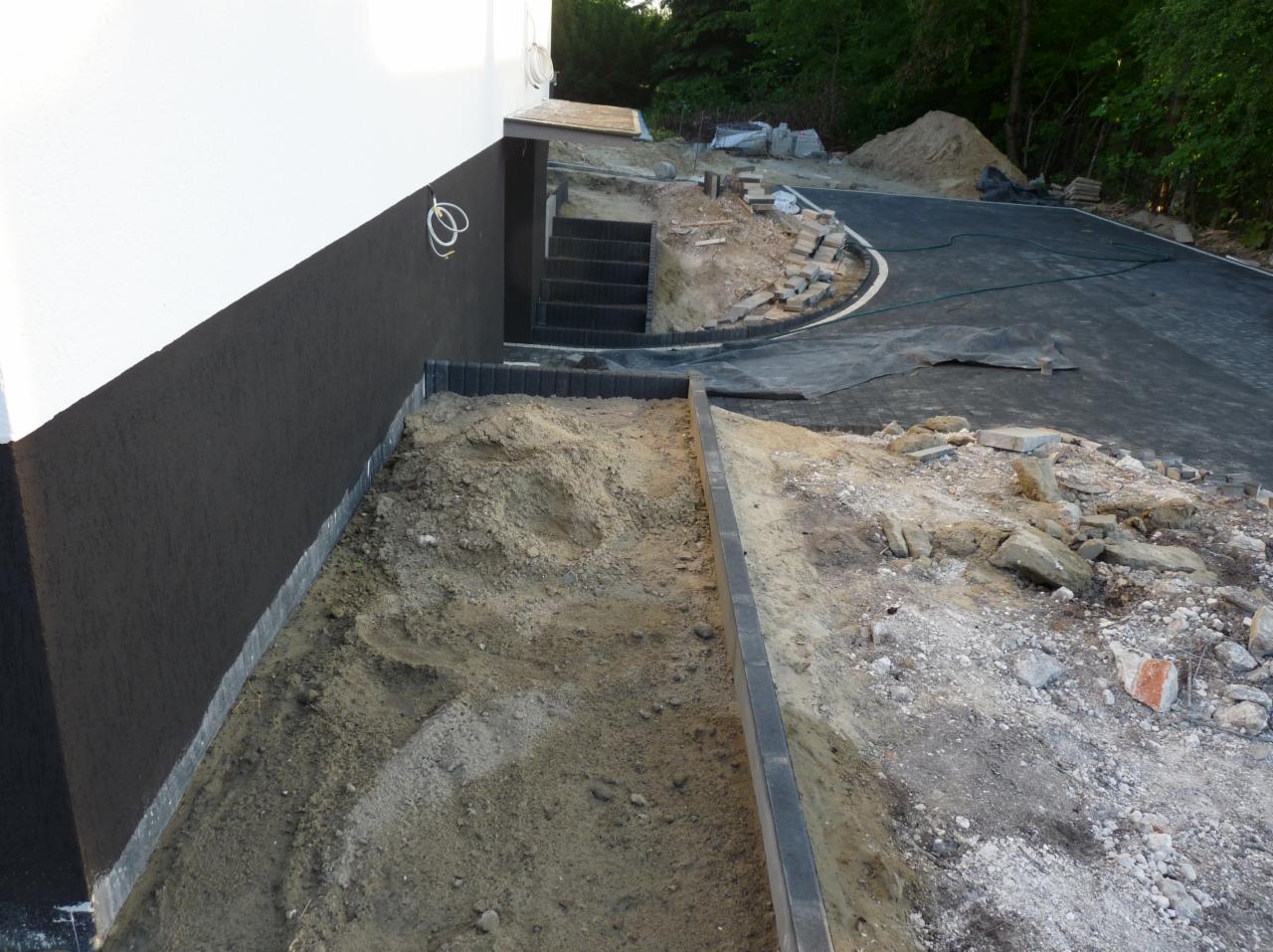 Przygotowanie terenu pod kostkę brukową obok budynku, widoczne krawężniki, piasek, gruz i ułożona nawierzchnia z kostki w tle.