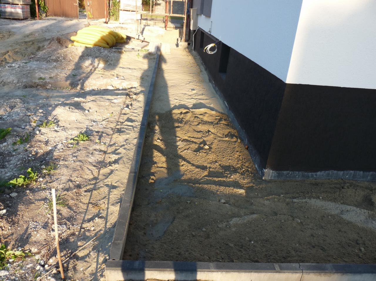 Przygotowanie terenu pod chodnik w Ornecie: widok na wykopane podłoże z ułożonymi obrzeżami wzdłuż fundamentu budynku i stos rur w tle.