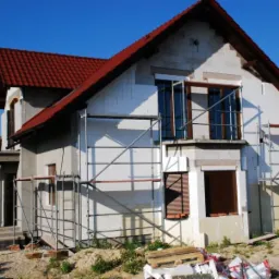 Dom jednorodzinny w trakcie ocieplania elewacji, widoczne rusztowania i warstwa izolacyjna na ścianach, czerwona dachówka.