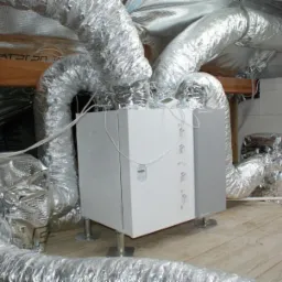 Centrala wentylacyjna z rekuperacją w przestrzeni poddasza, połączona z aluminiowymi przewodami wentylacyjnymi, widoczne elementy konstrukcji drewnianej i ściana z bloczków betonowych.