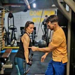 Bartłomiej Osuch - Kobieta ćwiczy na maszynie pod okiem trenera w siłowni. Trener wskazuje na jej biceps. W tle inne maszyny i napis na ścianie.