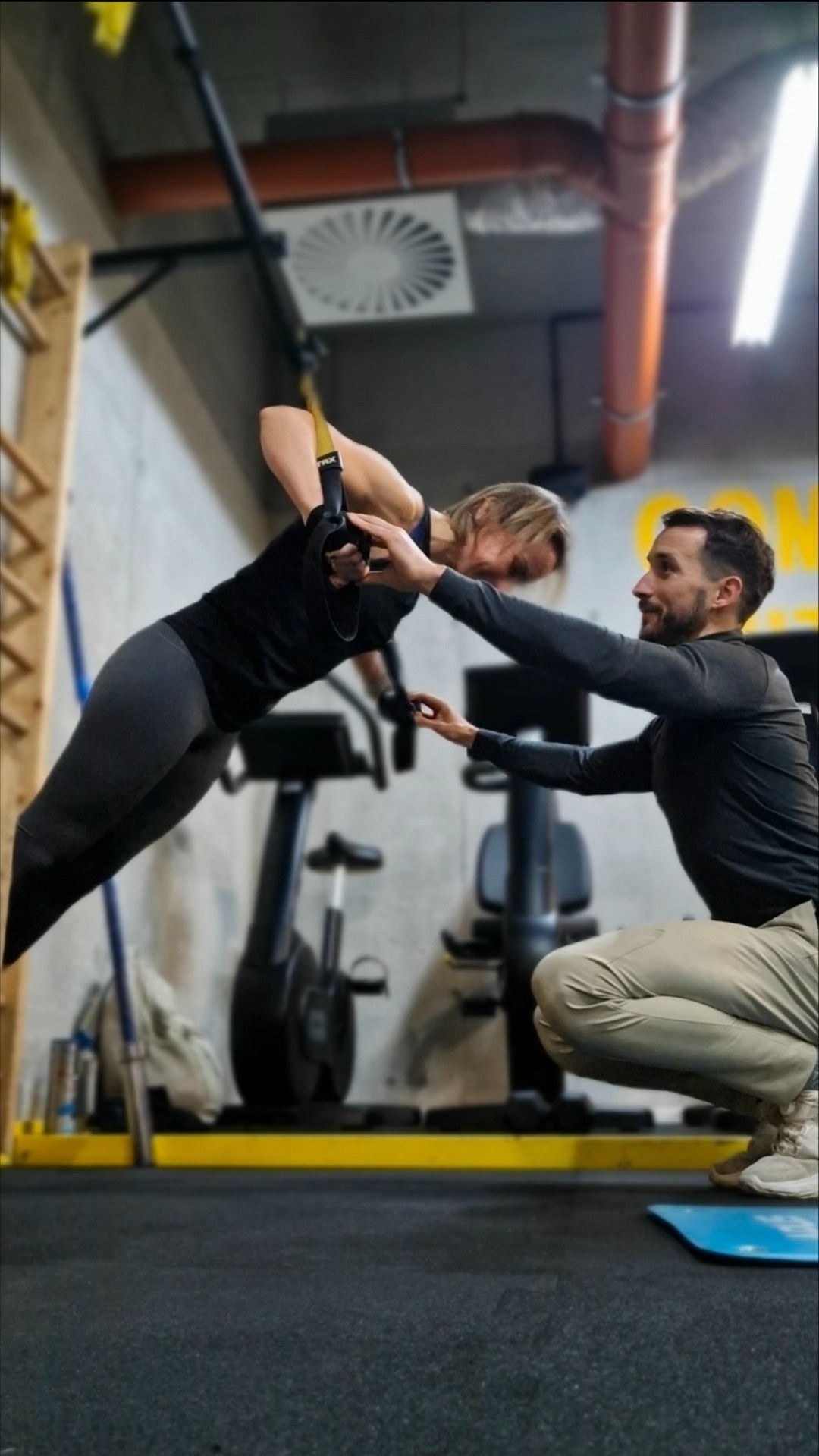 Kobieta ćwiczy na TRX pod okiem trenera w siłowni. Trener koryguje ustawienie dłoni. W tle drabinka gimnastyczna i rower stacjonarny. Trening funkcjonalny.