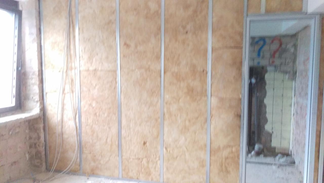 Ściana wyłożona wełną mineralną w trakcie montażu na metalowym stelażu, widoczne kable elektryczne przy oknie i nieukończone wykończenie w przejściu z namalowanymi znakami zapytania.