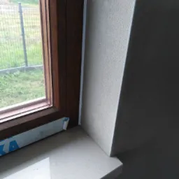 Fragment okna z drewnianą ramą, jasnym parapetem i fragmentem otynkowanej ściany w jasnych barwach; widoczny fragment folii ochronnej na ramie okiennej.