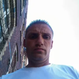 Autoportret mężczyzny w białej koszulce na tle ceglanego budynku z oknami, wykonany z dołu.