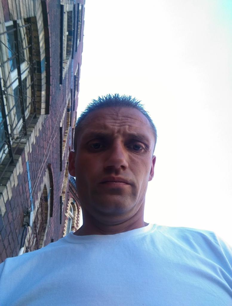 Autoportret mężczyzny w białej koszulce na tle ceglanego budynku z oknami, wykonany z dołu.