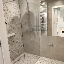 Nowoczesna kabina prysznicowa walk-in z szklaną ścianką, wykończona płytkami lastryko na ścianie i podłodze, minimalistyczny odpływ liniowy, szara armatura i grzejnik drabinkowy w tle.