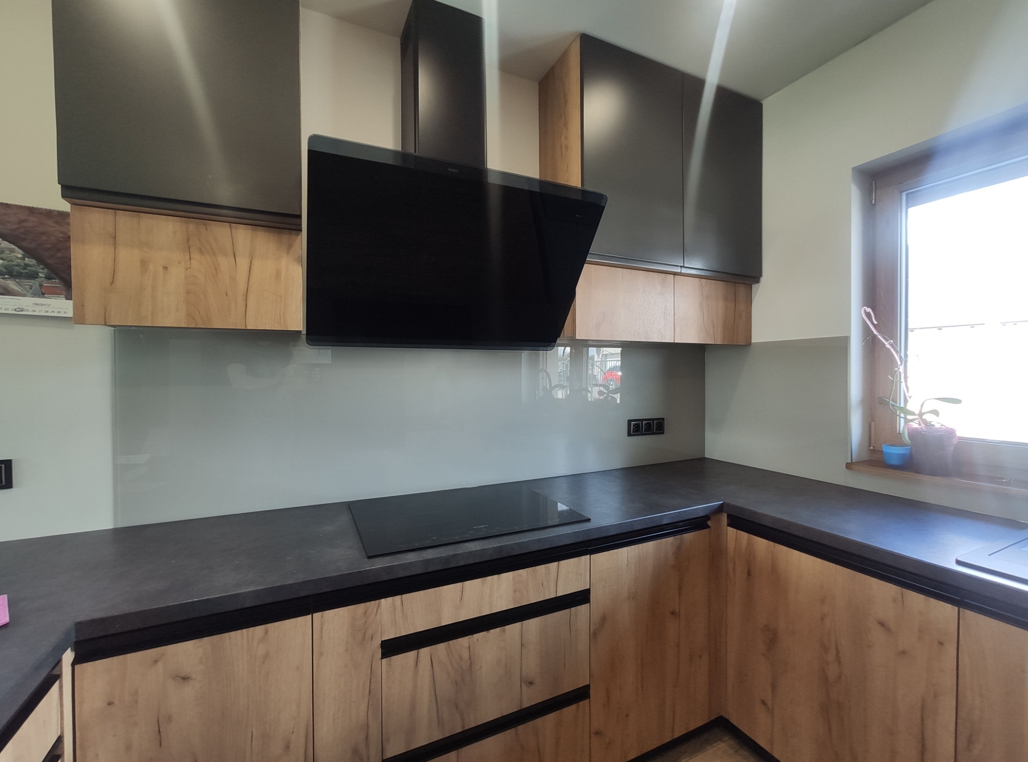 Nowoczesna kuchnia z drewnianymi frontami szafek, szarym blatem i szklanym panelem ściennym, czarny okap nad płytą indukcyjną, doniczka z rośliną na parapecie okna.