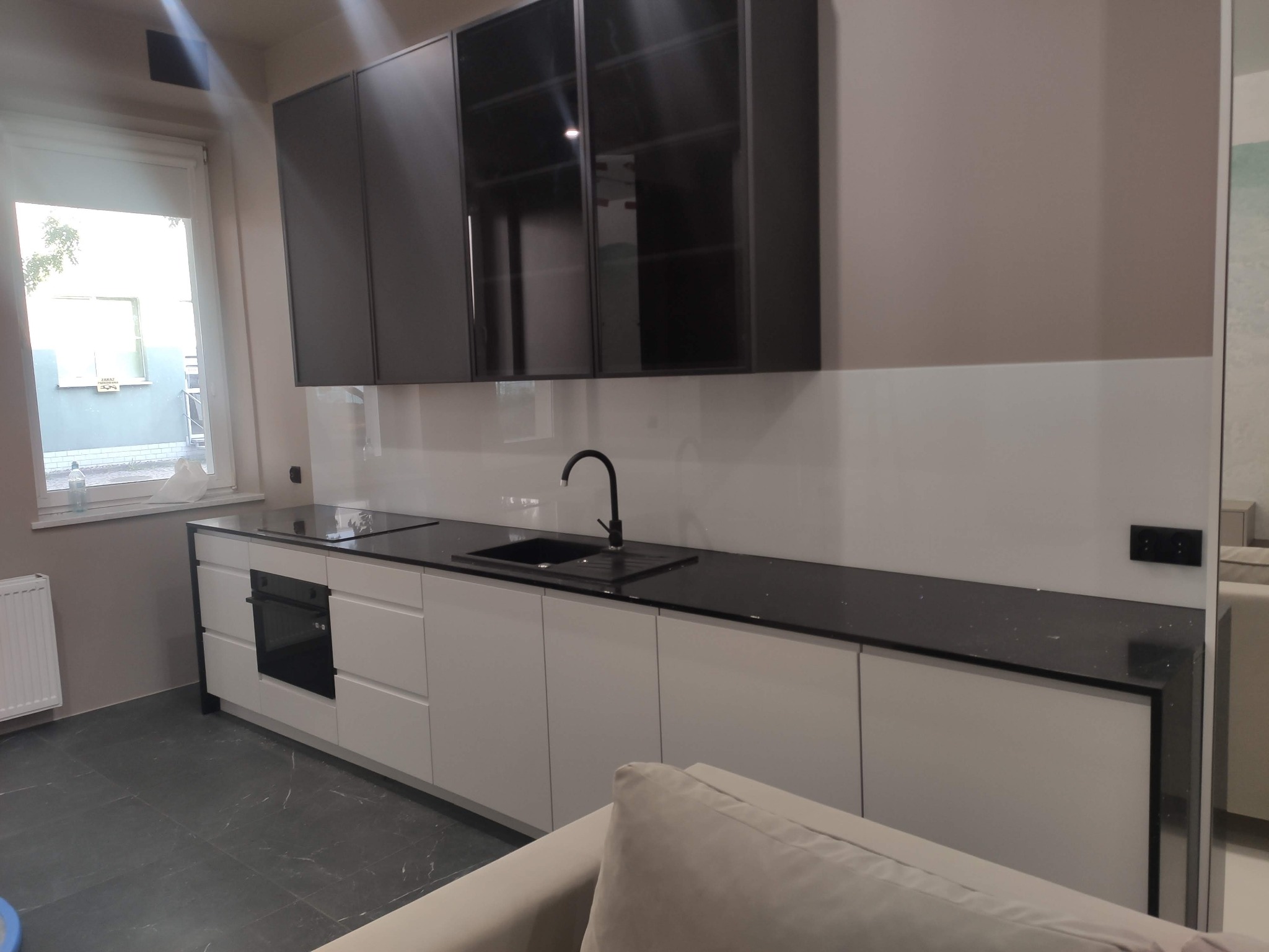 Nowoczesna kuchnia z białymi szafkami dolnymi i czarnymi szafkami górnymi, czarny blat z brokatem, czarna armatura, szara ściana i podłoga, widok z perspektywy salonu.