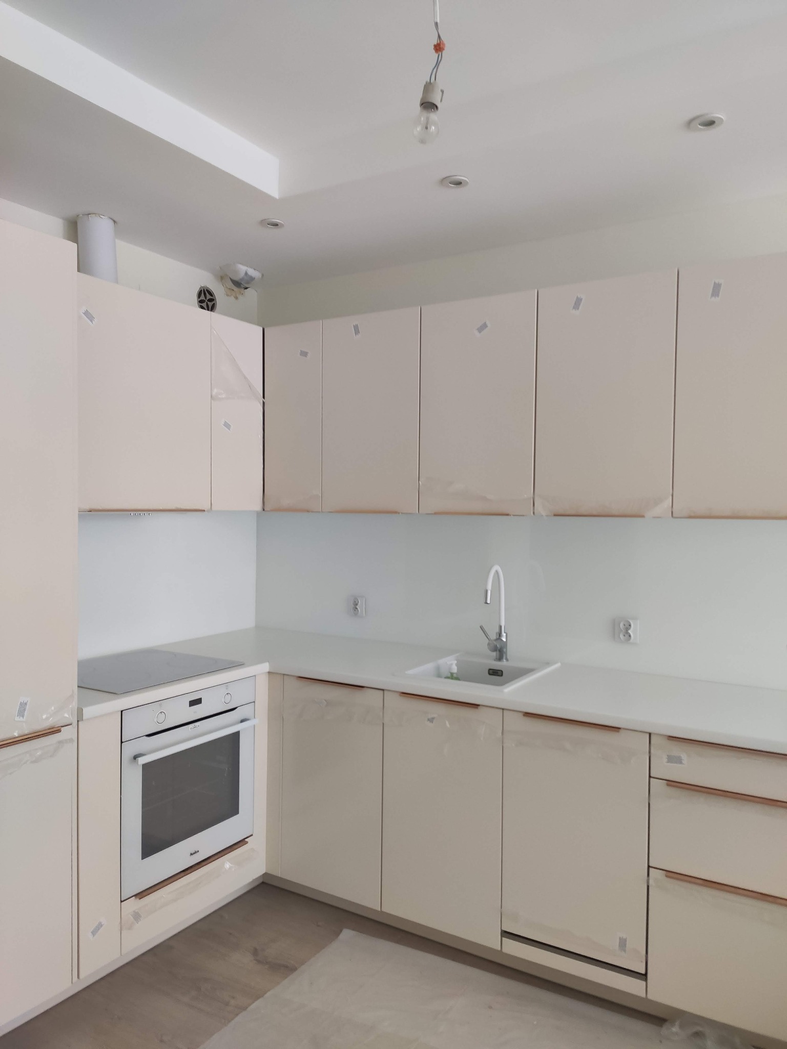 Jasna kuchnia z zamontowanymi szafkami, zabezpieczonymi folią ochronną, z widocznym zlewem, płytą indukcyjną i piekarnikiem, oraz wiszącą żarówką bez klosza.
