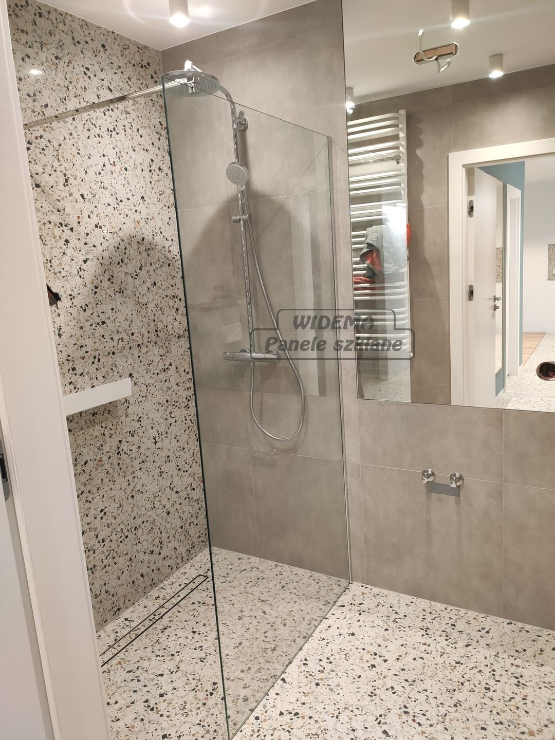 Nowoczesna kabina prysznicowa walk-in z szklaną ścianką, wykończona płytkami lastryko na ścianie i podłodze, minimalistyczny odpływ liniowy, szara armatura i grzejnik drabinkowy w tle.