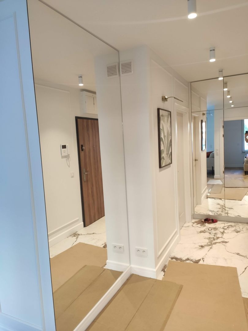 Korytarz z lustrzanymi ścianami, białe panele ścienne, marmurowa podłoga zabezpieczona kartonem, widoczne gniazdka elektryczne na ścianie.
