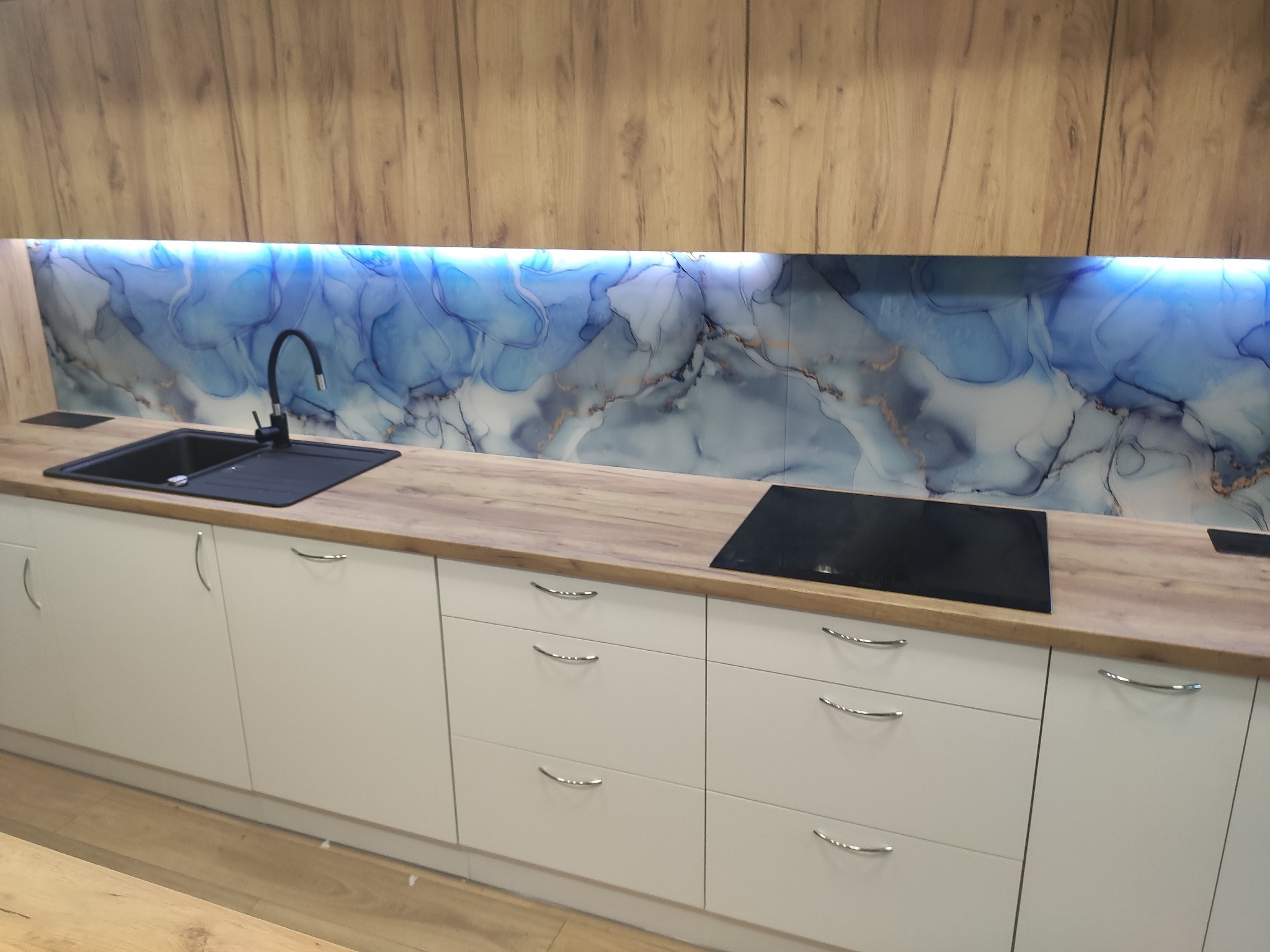 Nowoczesna kuchnia z białymi szafkami, drewnianym blatem i dekoracyjnym panelem ściennym z motywem marmuru w odcieniach błękitu i złota, oświetlonym od tyłu, z czarnym zlewem i płytą indukcyjną.