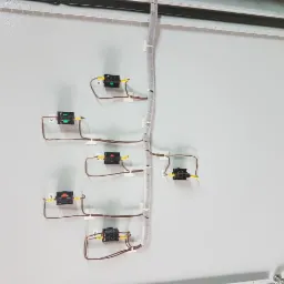 Wnętrze szarej szafy sterowniczej z zamontowanymi, czarnymi elementami elektrycznymi połączonymi brązowymi przewodami, zabezpieczonymi białymi opaskami i spiralą. Widoczne oznaczenia na żółtych...
