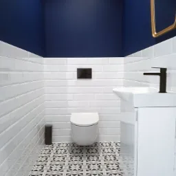 Nowoczesna łazienka z białymi, prostokątnymi płytkami ściennymi, granatową g&oacute;rną częścią ścian, wzorzystą podłogą, wiszącą toaletą i białą umywalką z czarną baterią.