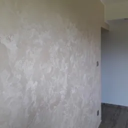 Ściana pokryta dekoracyjną, jasną gładzią w stylu weneckim, z widoczną fakturą i subtelnym połyskiem, oświetlona naturalnym światłem w rogu pomieszczenia.