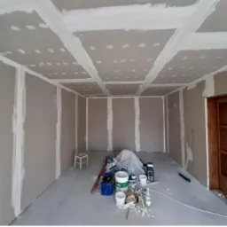 Pokój w trakcie remontu z zamontowanymi płytami gipsowo-kartonowymi na ścianach i suficie, widoczne łączenia płyt wypełnione masą szpachlową, narzędzia i materiały budowlane na podłodze.