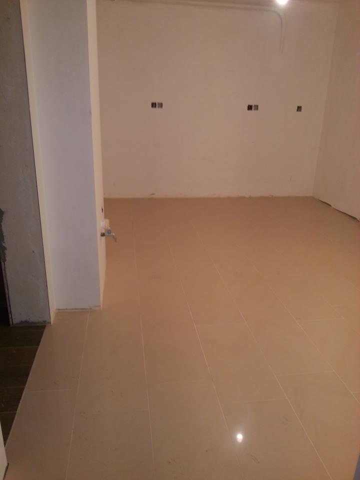 Beżowe płytki podłogowe ułożone w pustym pomieszczeniu, widoczne puszki instalacyjne na ścianie, fragment kolumny z wystającą instalacją wodną.