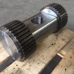 WR Rafał Wisiński - Cylindryczny, metalowy element z ząbkowanymi kołnierzami po obu stronach, leżący na drewnianej palecie. Widoczne otwory i śruby mocujące.