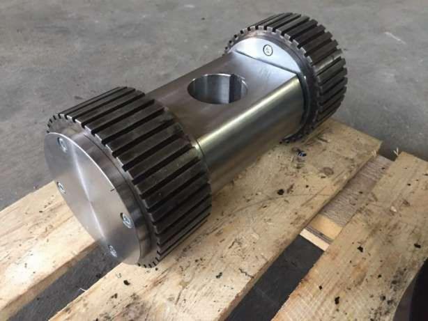 Cylindryczny, metalowy element z ząbkowanymi kołnierzami po obu stronach, leżący na drewnianej palecie. Widoczne otwory i śruby mocujące.