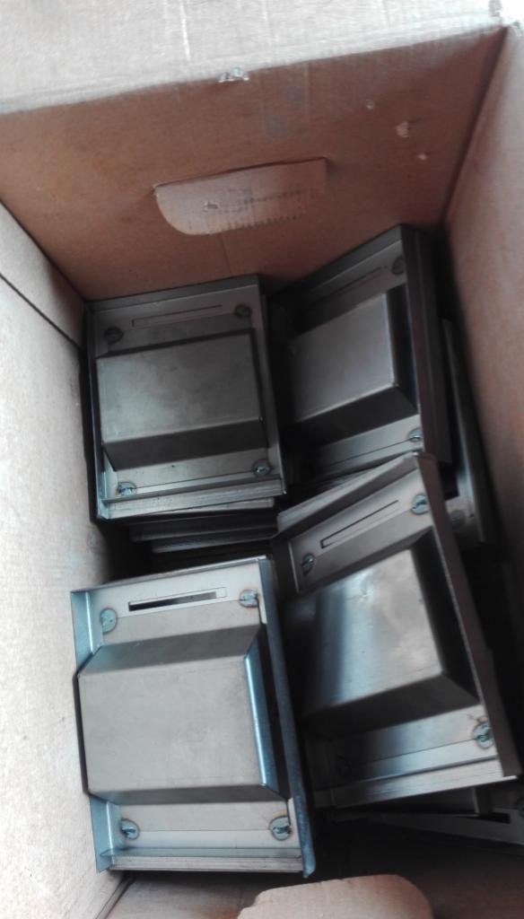 Metalowe obudowy z otworami na śruby, ułożone w kartonowym pudle, widoczne ślady montażu.