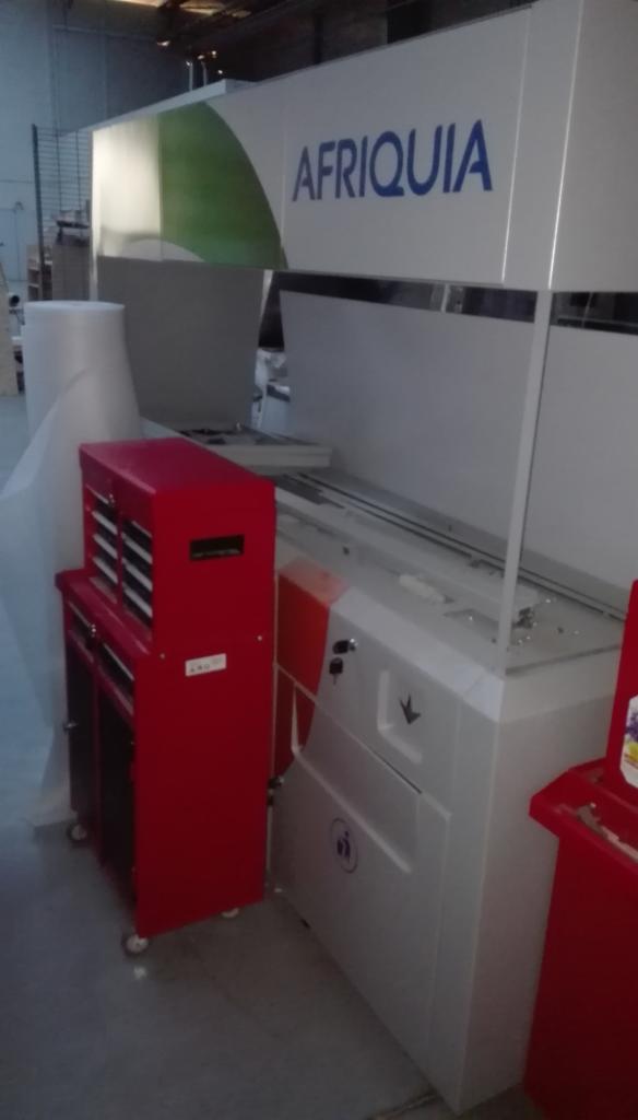 Duża, biała maszyna przemysłowa z napisem AFRIQUIA, obok czerwona szafka narzędziowa na kółkach, w tle widoczny magazyn.