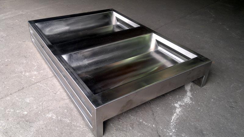 Dwu-komorowa, spawana konstrukcja ze stali nierdzewnej, prawdopodobnie taca lub pojemnik, na niskich nóżkach, leżąca na betonowej podłodze.