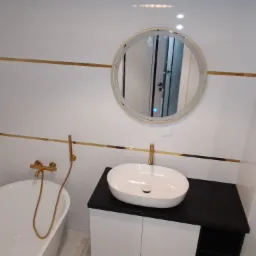 Nowoczesna łazienka z białymi płytkami, złotymi akcentami, okrągłym lustrem z podświetleniem LED, białą umywalką nablatową i złotą armaturą.