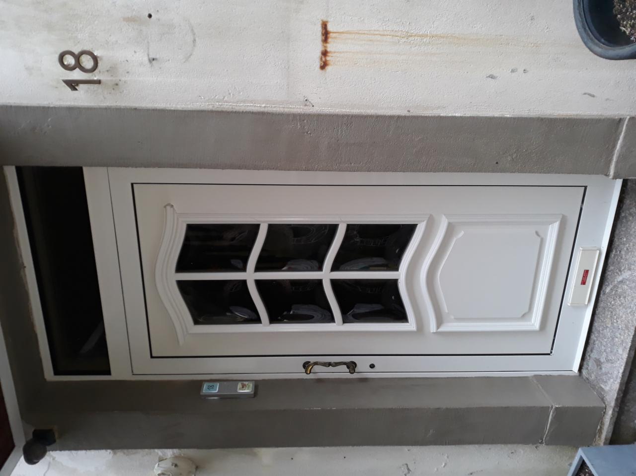 Białe okno piwniczne z ozdobnymi szprosami i widocznym numerem budynku '18' na ścianie powyżej.