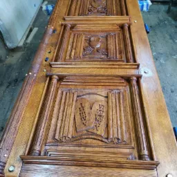 Odrestaurowane, drewniane drzwi z rzeźbionymi detalami, ukazujące motywy religijne, leżące poziomo na niebieskich podkładkach w warsztacie.