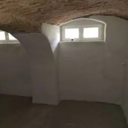 Surowe wnętrze piwnicy z łukowym, ceglanym sklepieniem, otynkowanymi ścianami z małymi oknami i betonową podłogą w odcieniach szarości.