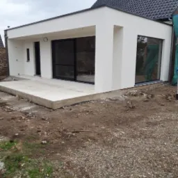 Nowoczesna, biała dobudówka do domu z dużymi, przesuwnymi oknami i betonowym tarasem w trakcie budowy, widok z zewnątrz na nierówny teren.