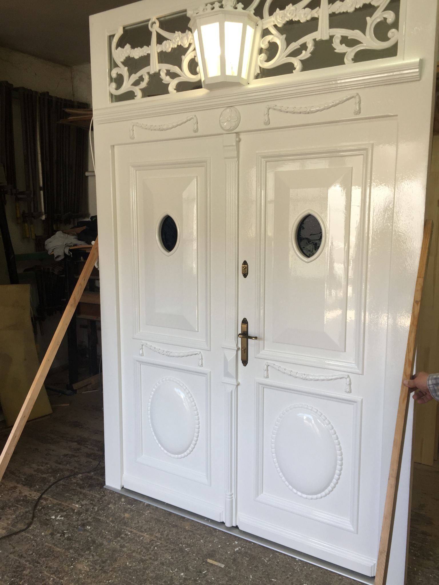 Białe, dwuskrzydłowe drzwi z dekoracyjnymi elementami i owalnymi przeszkleniami, oświetlone lampą w stylu retro, wsparte drewnianymi listwami w warsztacie.