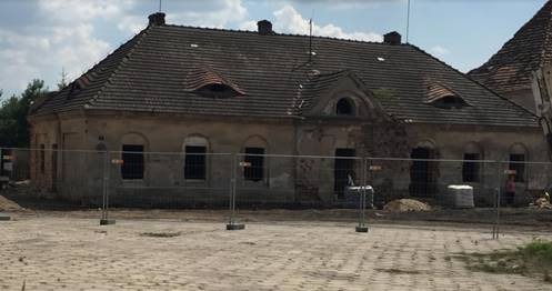 Stary, zaniedbany budynek z elementami architektury zabytkowej, zabezpieczony ogrodzeniem tymczasowym, ukazujący postęp prac remontowych i częściowo odrestaurowaną elewację z widocznymi ubytkami...