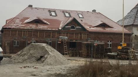 Ceglany budynek w trakcie prac elewacyjnych, widoczna sterta piasku, rusztowania oraz palety z materiałami budowlanymi, lekko ośnieżony dach i otoczenie, pochmurny dzień.