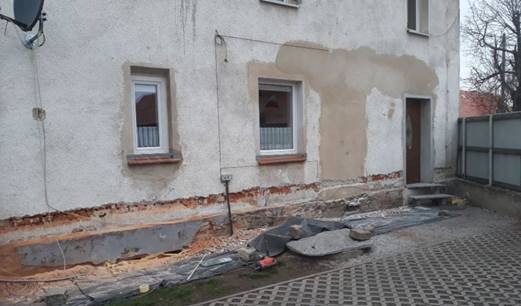 Przygotowania do prac elewacyjnych: odsłonięty fundament budynku z widocznymi cegłami, częściowo otynkowana ściana z dwoma oknami i wejściem, zabezpieczenia folią i gruzem na podłożu.